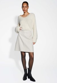 Femme portant un pull en laine blanche moelleux, une jupe beige clair, des collants noirs transparents et des bottines noires, se tenant devant un fond blanc uni.