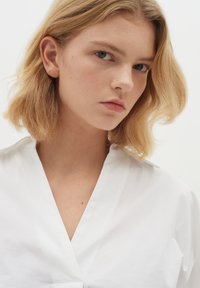White collared shirt with a V-neck, featuring smooth texture and minimalist design. Model displays short, textured blonde hair.