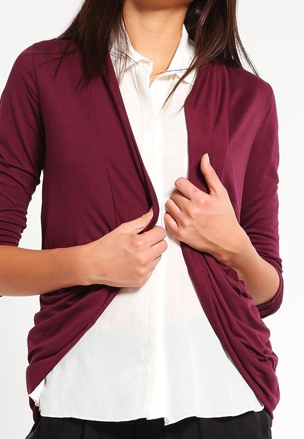 Personne ajustant un cardigan bordeaux à manches longues sur une chemise blanche boutonnée, visible des épaules à la taille.