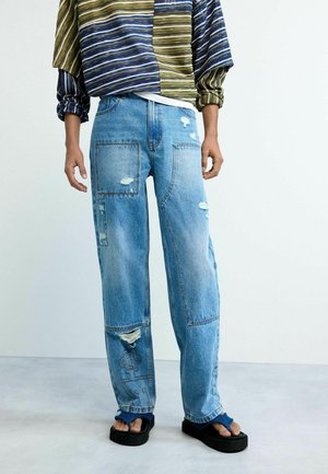 Person wearing distressed patchwork blue jeans, striped long-sleeve top, and black platform sandals with blue straps, standing on gray floor.