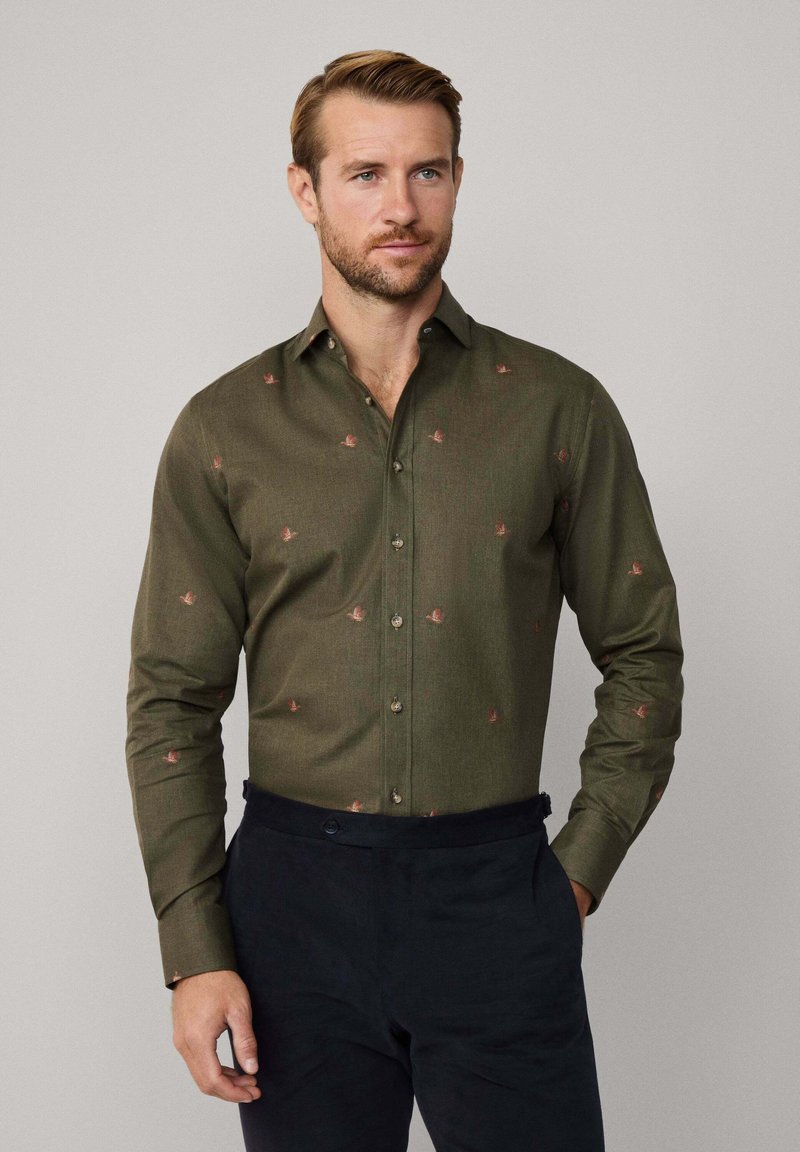 Man in een olijfgroen overhemd met korte mouwen, met kleine rode vogelpatronen, in donkere marineblauwe broek, staande tegen een effen achtergrond.