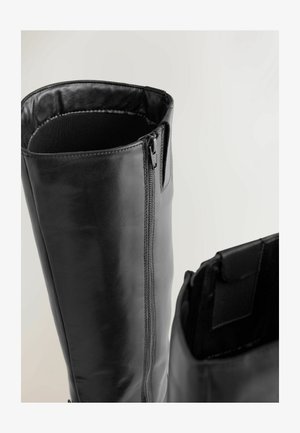 Cizme negre din piele până la genunchi, cu suprafață netedă, fermoar lateral și căptușeală interioară moale. Au vârf rotunjit și accente minimaliste.