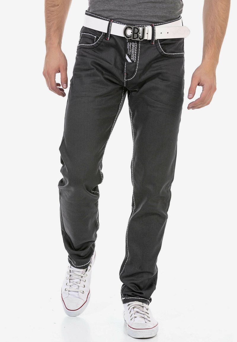 Zwarte denim jeans met witte stiksels, slim fit, vijf zakken en een witte riem. Draagt wit met rode accenten sportschoenen.