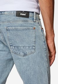 Los pantalones vaqueros de denim azul claro presentan un corte recto, bolsillos traseros y una etiqueta negra en la cintura con texto blanco. Hay detalles de costura visibles.