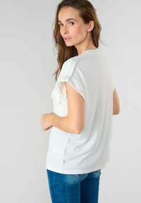 Top bianco a maniche corte con una vestibilità ampia, caratterizzato da una tasca laterale e dettagli sulle spalle. Abbinato a jeans blu, texture del tessuto liscia.