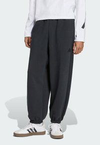 Pantalons de survêtement gris foncé avec une taille �élastique et des poignets, arborant un logo discret sur le côté. Associés à des baskets blanches avec des accents noirs.