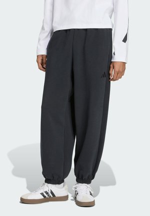 Pantalons de survêtement gris foncé avec une taille élastique et des poignets, arborant un logo discret sur le côté. Associés à des baskets blanches avec des accents noirs.
