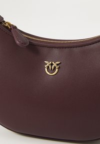 Burgundy színű bőr kézitáska sima felülettel, arany tónusú logóval levél motívumokkal és cipzáras záródással a tetején.
