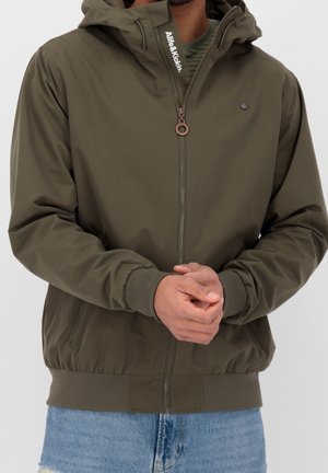 Man draagt een olijfkleurig ritsvest met capuchon, voorzien van een logo op de ritssluitingflap en geribbelde manchetten, gecombineerd met een blauwe spijkerbroek, handen gevouwen.