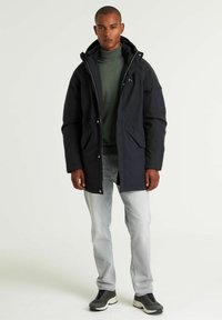 Tamno morska parka s patentnim zatvaračem i kopčom, kapuljačom i više džepova; kombinirana s zelenim puloverom s visokim ovratnikom i svijetlim traperskim hlačama.