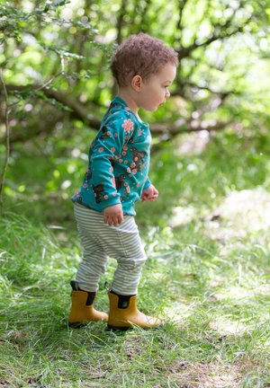 Kind, der ein türkisfarbenes Langarmshirt mit Blumen- und Tiermotiven, gestreiften grauen Leggings und gelben Gummistiefeln trägt, läuft auf Gras.