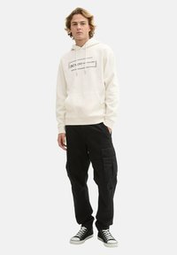 Crème hoodie met grafische print aan de voorkant en trekkoorden, gecombineerd met zwarte cargo-broeken met zakken, afgewerkt met zwarte sneakers.