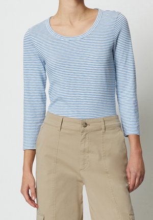 Personne portant un haut à manches longues rayé bleu clair et blanc, associé à un pantalon beige taille haute, sur un fond uni.