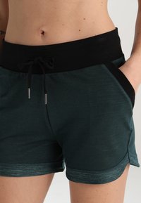 Detailní záběr na osobu v tmavě zelených kraťasech s černým elastickým pasem a šňůrkou, ruka v kapse, ukazující holé břicho.