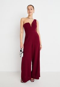 Jumpsuit bordeaux con una spalla, scollatura a V, vestibilità su misura e gambe larghe. La modella tiene in mano una piccola borsa scintillante e indossa gioielli argentati.