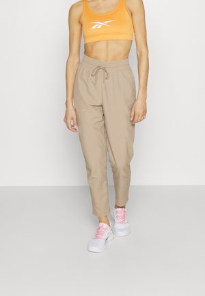 Frau tr trägt einen orangefarbenen Sport-BH, beige Zugbandsporthosen und weiße Sneaker mit rosa Schnürsenkeln, stehend vor einem schlichten weißen Hintergrund.