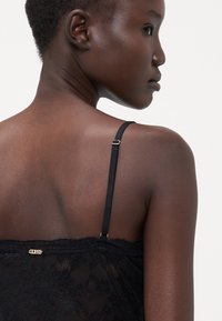 Camisole en dentelle noire avec des bretelles fines réglables, présentant un ourlet festonné et un détail en métal doré décoratif au dos.