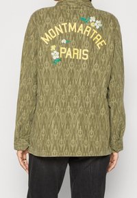 Olijfgroene jas met een ikatpatroon, voorzien van bloemenborduursel en "MONTMARTRE PARIS" in het geel op de rug. Relaxte pasvorm en lange mouwen.
