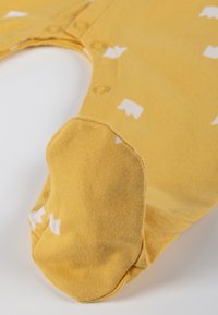 Body per neonati in cotone giallo con motivi astratti bianchi, bottoni a pressione sul collo e piedi coperti per mantenere il calore. Texture morbida.