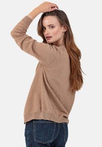 Femme aux longs cheveux bruns portant un pull marron et un jean bleu, levant une main pour toucher sa tête, regardant par-dessus son épaule.