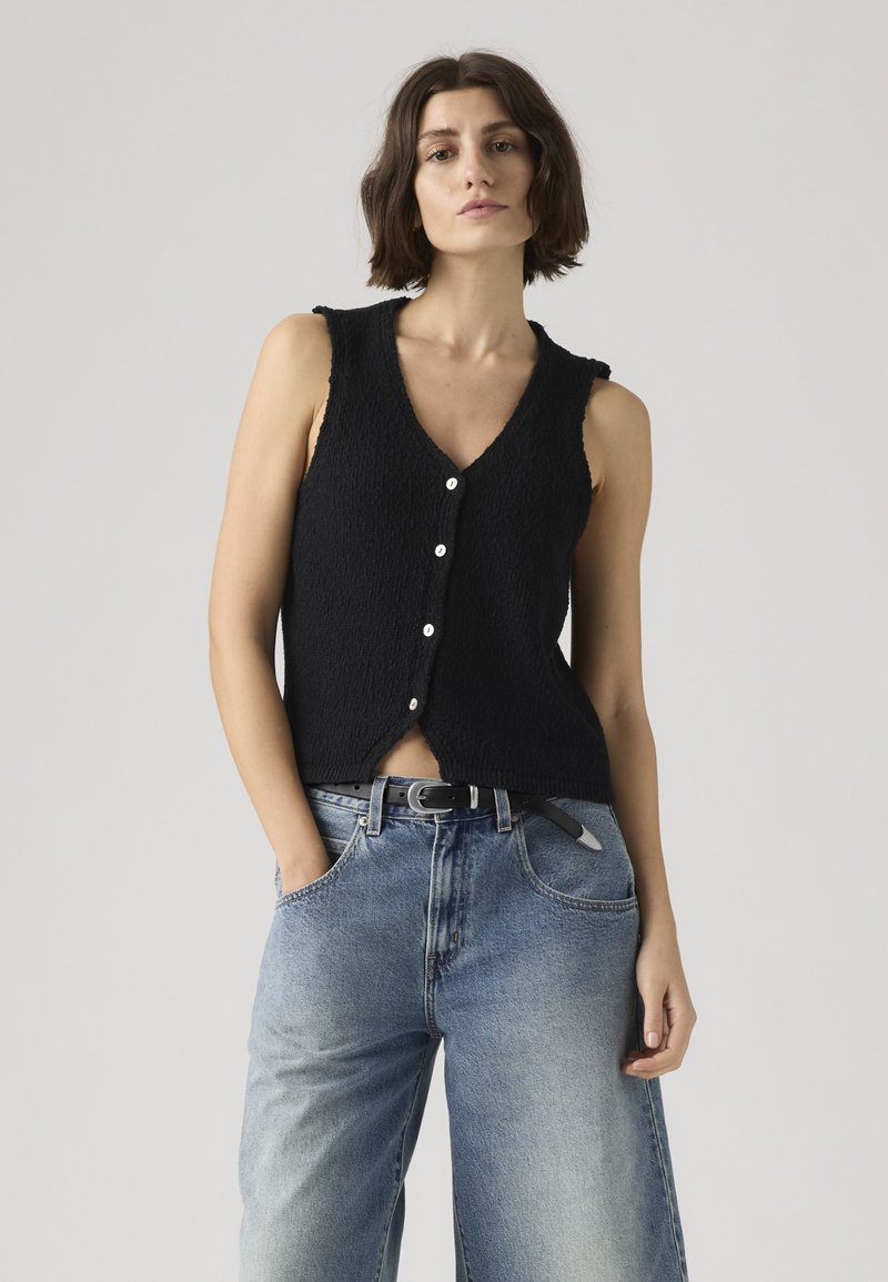 Woman with short dark hair wearing a sleeveless black buttoned vest and loose blue jeans with a black belt, standing with one hand in pocket.