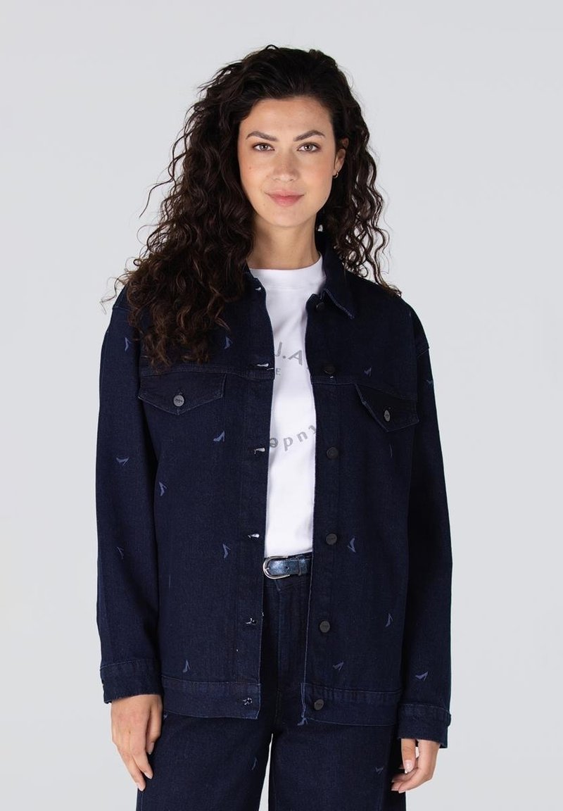 Donkerdenimjack met twee borstzakken en een knoopsluiting, voorzien van een subtiel patroon van kleine blauwe vormen. Gedragen over een wit shirt.