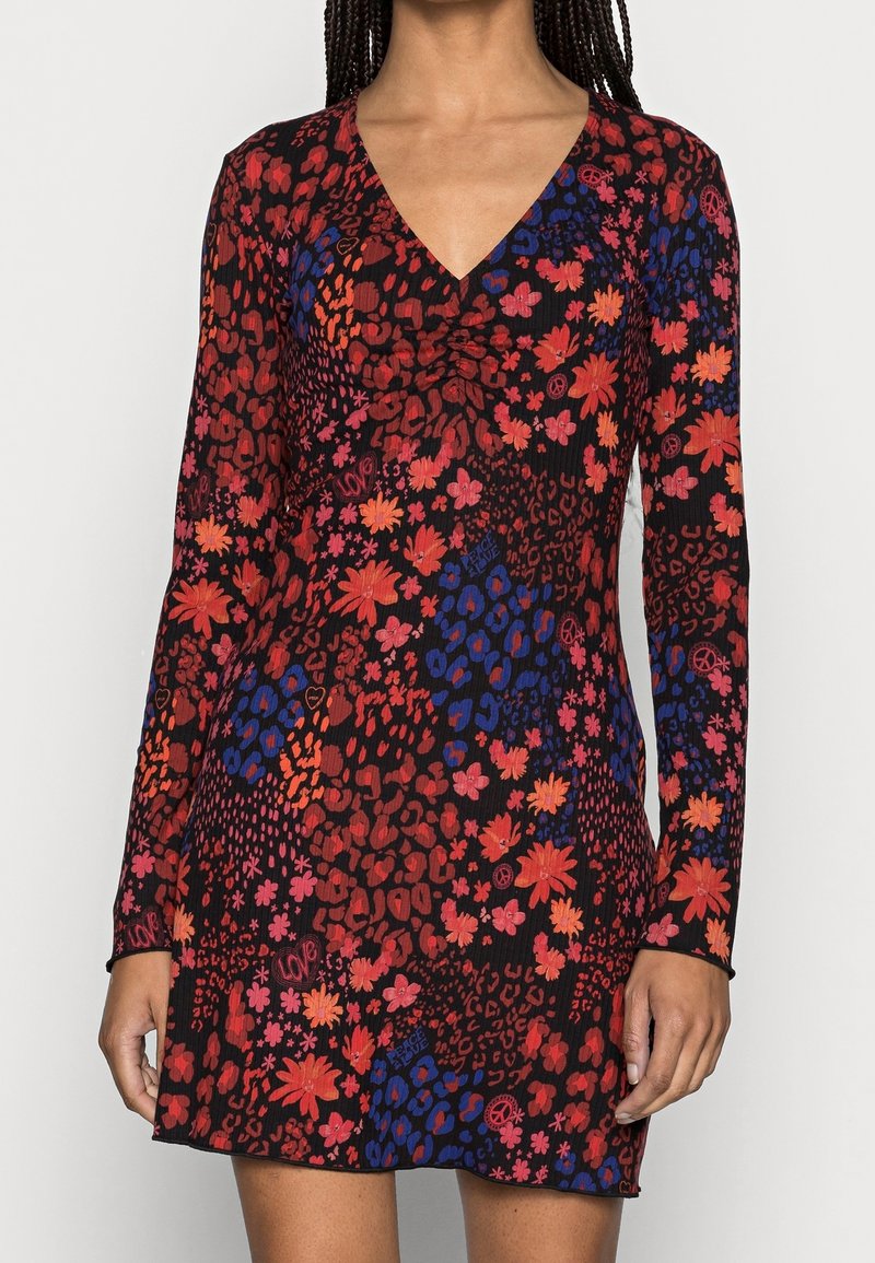Femme portant une robe noire à manches longues avec des motifs floraux et animaliers rouges, orange, roses et bleus et un col en V.