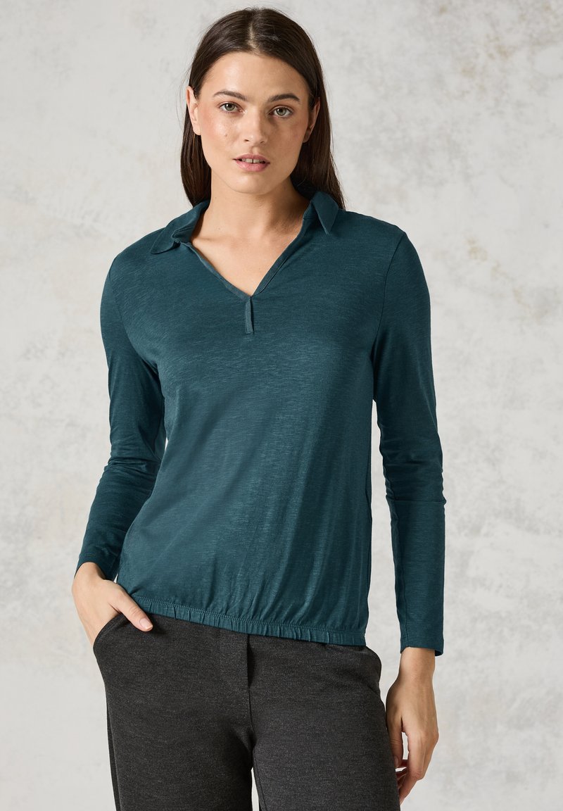 Teal Langarmshirt mit knopflosem Verschluss, Kragen und gerafftem Saum. Weicher, leichter Stoff mit leichtem Glanz. Getragen mit dunklen Hosen.