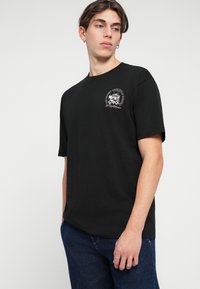 T-shirt preta de manga curta feita de algodão. Apresenta uma estampagem gráfica no lado esquerdo do peito e um decote redondo. Textura suave.