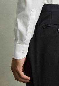 Weißes Baumwollhemd mit Ärmelaufschlag und zwei runden Knöpfen, mit gerader Kante, kombiniert mit dunklen, taillierten Hosen mit einer einzelnen Gesäßtasche.