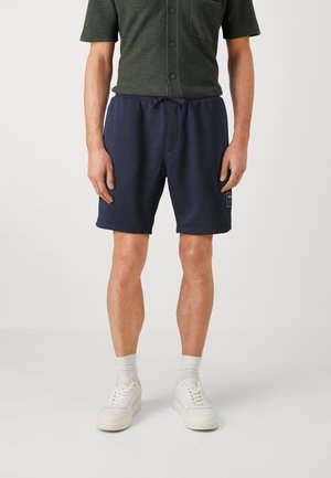 Homme portant une chemise vert foncé à manches courtes, un short bleu marine avec cordon de serrage et poche latérale, des chaussettes blanches et des baskets blanches, debout sur un fond uni.