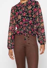 Blusa floreale con maniche lunghe, base nera, fiori rosa e verdi vivaci, vita stretta, abbinata a pantaloni marroni a coste.