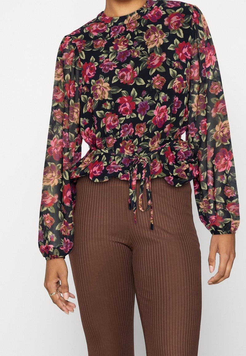 Blusa floreale con maniche lunghe, base nera, fiori rosa e verdi vivaci, vita stretta, abbinata a pantaloni marroni a coste.