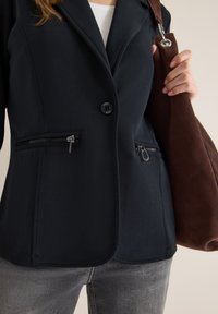 Marineblauwe blazer van zacht materiaal, met een enkele knoopsluiting, ritszakken en een zijschuif. Gecombineerd met grijze jeans en een bruine tas.