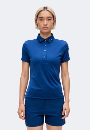 Junge Person mit kurzen blonden Haaren, die ein blaues Polohemd mit kurzen Ärmeln und passende blaue Shorts trägt, steht mit Blick nach vorne.
