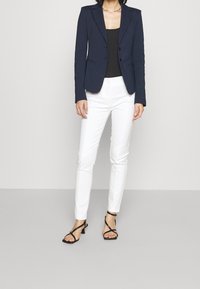 Granatowa marynarka z wciętymi klapami, połączona z czarnym topem na ramiączkach i białymi dopasowanymi spodniami, uzupełniona czarnymi sandałami na paski.