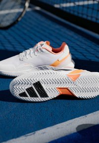 Λευκά αθλητικά παπούτσια με πορτοκαλί λεπτομέρειες, διαθέτοντας κλωστοϋφαντουργικό επάνω μέρος με υφή, αντίθετη μαύρη σόλα και στρογγυλεμένο σχέδιο για αθλητική απόδοση.