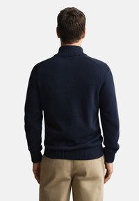 Temno modro pleten pulover z visokim ovratnikom, rebrastimi manšetami in robom. Gladek tekstura in oprijeta zasnova z detajli na šivih ramen.
