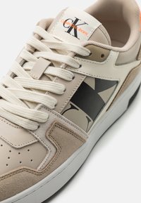 Baskets beiges avec une tige en cuir texturé et en daim, présentant des accents de logo noirs, des lacets beiges et une semelle en caoutchouc blanche.