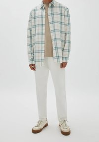 Chemise à carreaux blancs avec des lignes bleues et jaunes, portée sur un t-shirt beige. Associée à un pantalon blanc et des baskets crème avec des semelles marron.