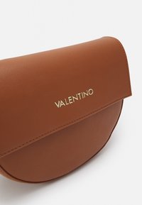 Brun läderhandväska med rundad form, med ett klaffstängning och guld "Valentino"-logotyp. Slät textur och synliga sömdetaljer.