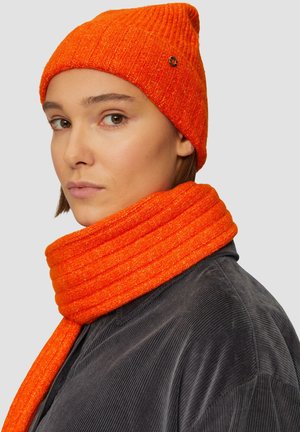 Oranje geribbelde gebreide beanie en bijpassende sjaal, zachte textuur, met een zwart accentlogo op de beanie. Draagt over een donker korduroy overhemd.