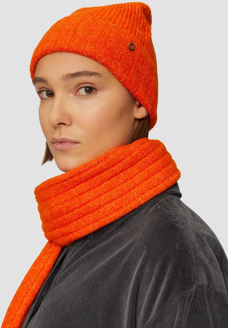 Oranje geribbelde gebreide beanie en bijpassende sjaal, zachte textuur, met een zwart accentlogo op de beanie. Draagt over een donker korduroy overhemd.