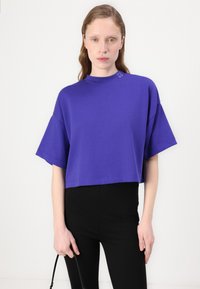 Maglietta viola cropped con scollatura alta e maniche corte oversize, con sottile logo "HUGO" vicino al colletto. Abbinata a pantaloni neri.