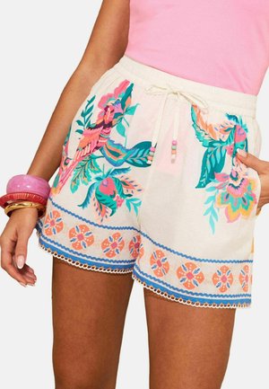 Nainen pukeutuneena valkoisiin shortseihin, joissa on värikäs kukka- ja lintukuosi, kiristysnyöri helminauhalla vyötäröllä, yhdistettynä vaaleanpunaiseen toppiin ja paksuihin rannekoruihin.