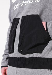 Šedá mikina s kapucí, která má černou náprsní kapsu se zapínáním na patentky, elastický pas a potisk na přední straně. Tkanina vypadá texturovaně.