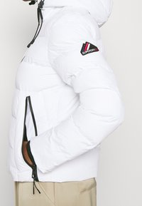 Hvid puffjakke med hætte, med sorte lynlåse, elastiske manchetter og et rød-sorte logopatch på venstre ærme. Glat tekstur.