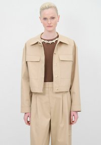 Giacca cropped beige chiaro con due tasche frontali, bottoni beige e colletto. Indossata sopra un top marrone e pantaloni a vita alta coordinati.