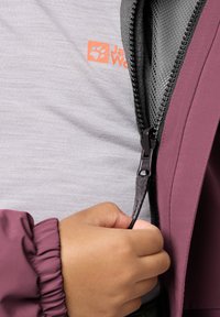 Hand zipping up a dark purple jacket over a light gray shirt with orange logo, showing fabric texture and zipper detail.