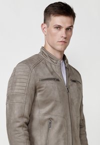 Jachetă gri din suede, având un guler înalt, două buzunare pentru piept cu fermoar, detalii cu nervuri pe umeri și o textură netedă.